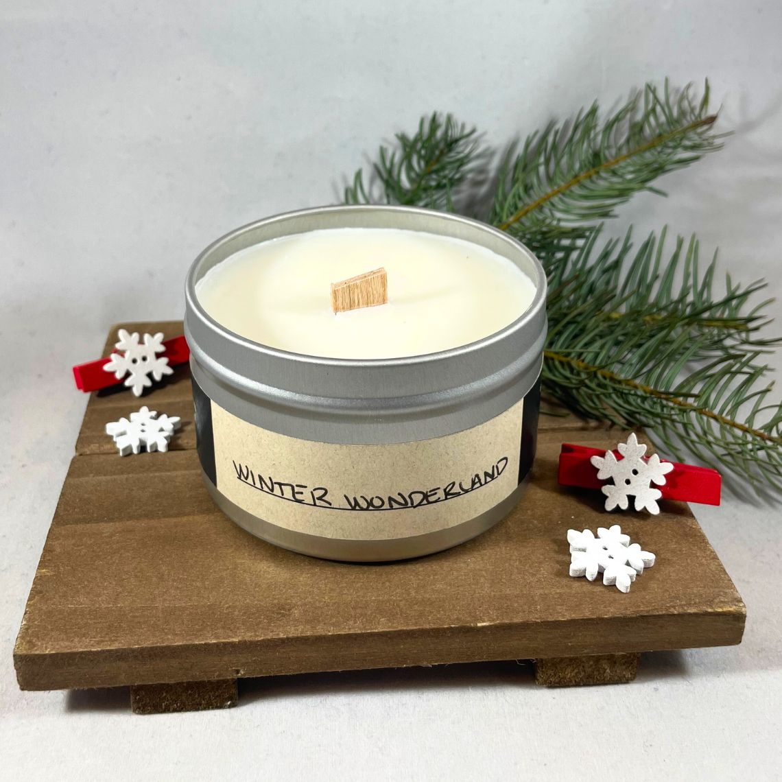 Candle in a tin labeled 'Winter Wonderland' with decorative snowflakes and a branch on a wooden stand.